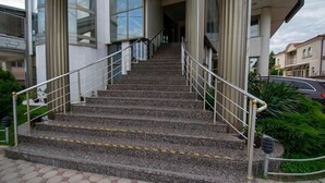 Degraus de pedra ao ar livre que levam à entrada de um edifício, com guarda-corpos de metal e um céu claro ao fundo.