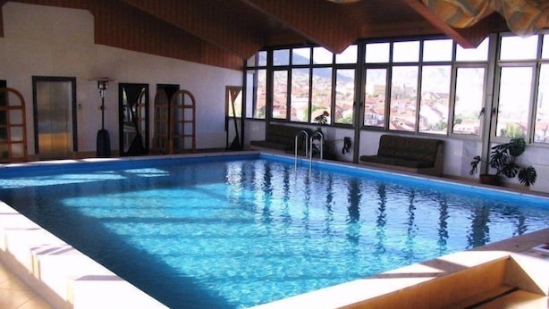 Piscina interna com água azul cristalina, deck de madeira e janelas amplas com vista para o exterior.