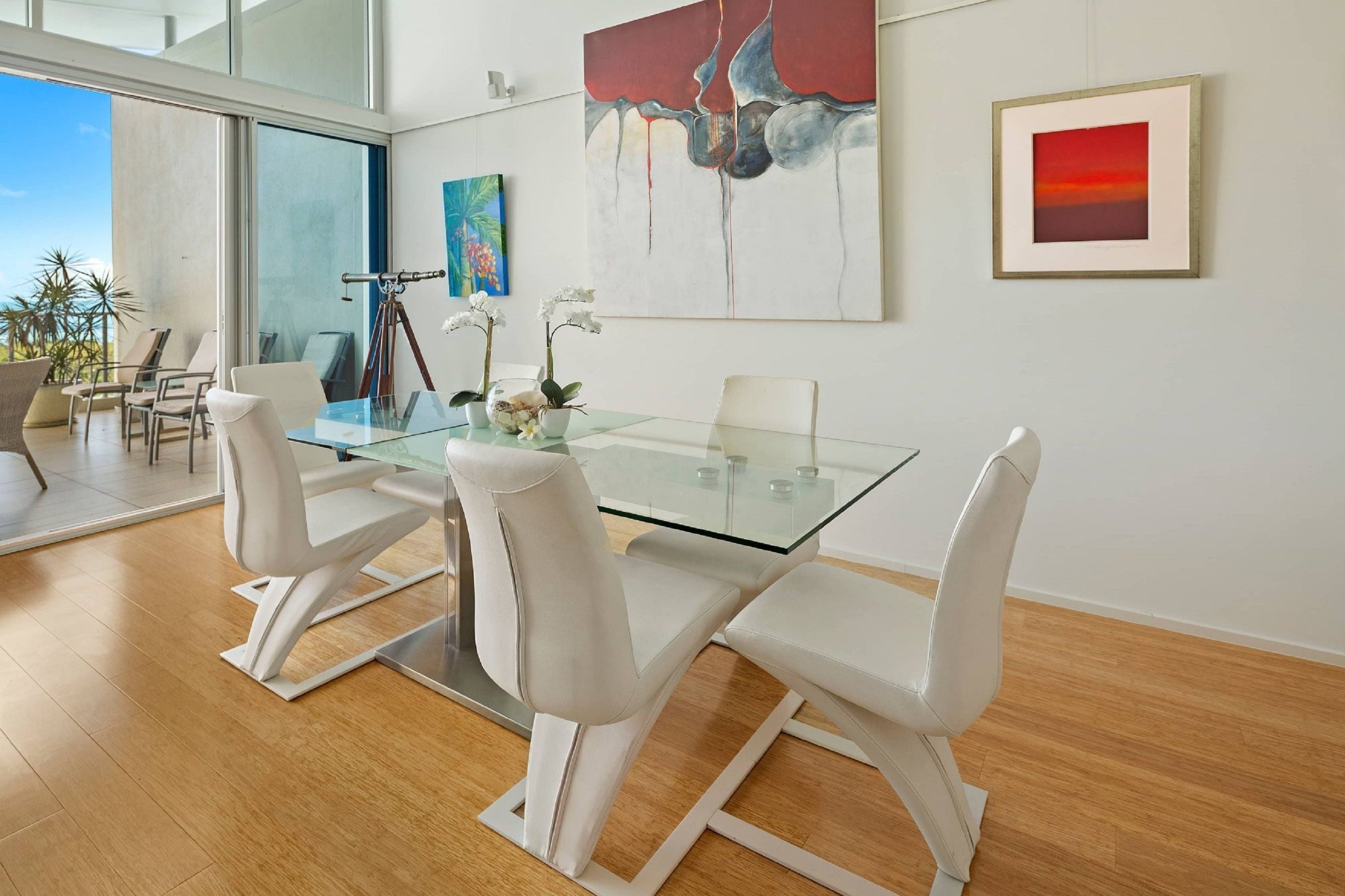 A modern dining area with a glass table and white chairs, a large abstract painting, and a framed picture on the wall.
