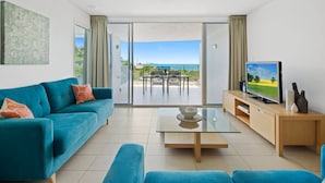 Een moderne woonkamer met een blauwgroene bank, een glazen salontafel, een flatscreen televisie en een schuifpui naar een balkon met uitzicht op de zee.