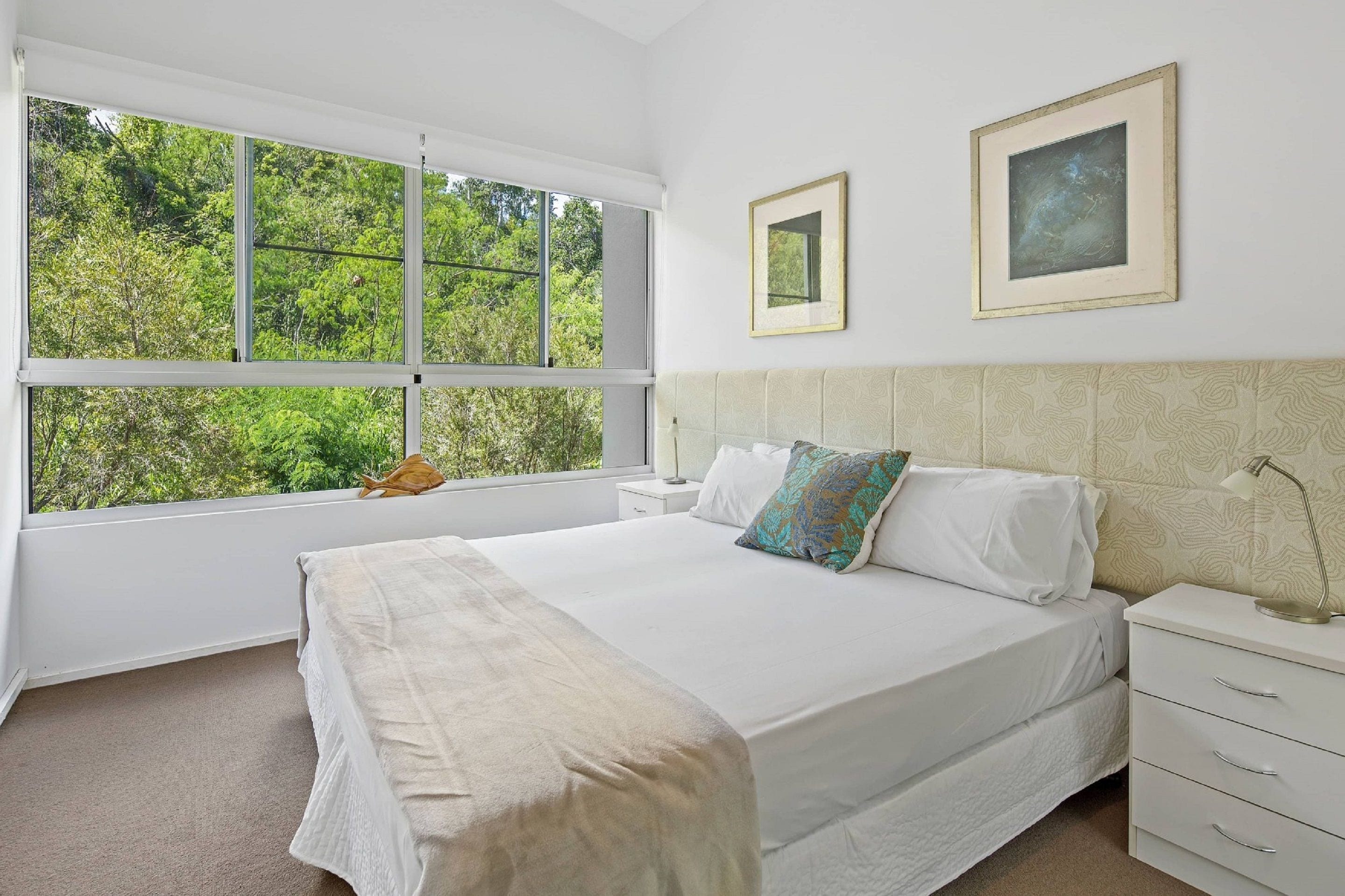 A bedroom with a large window, a bed, a nightstand, and two framed pictures on the wall.