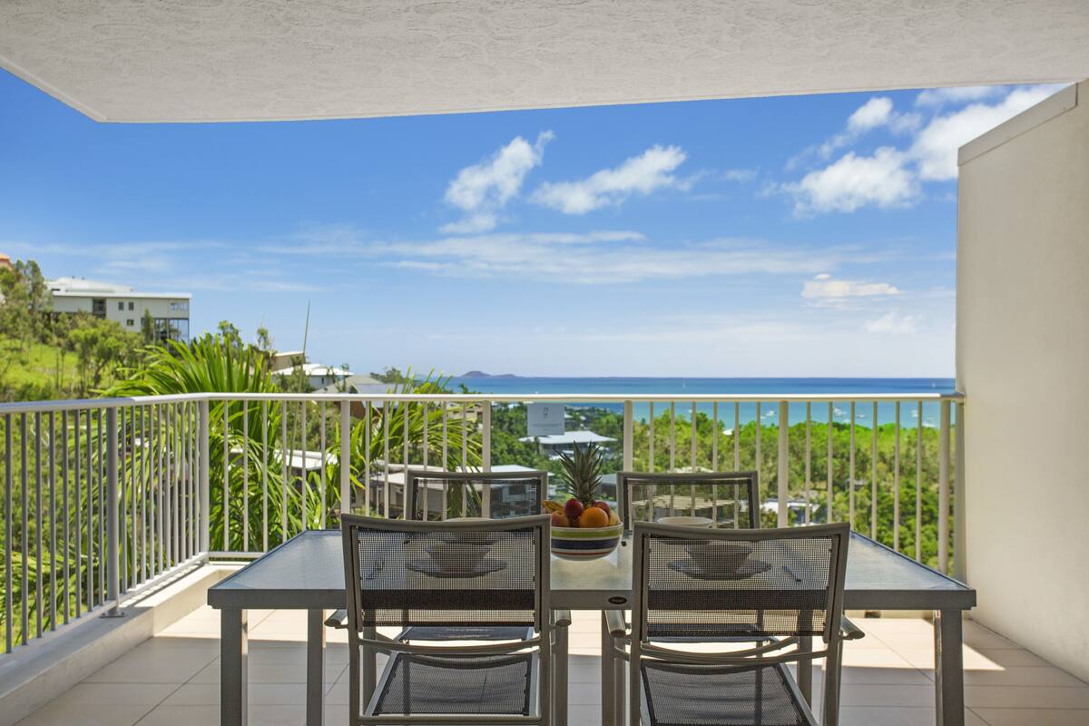 Un balcon avec une table et des chaises, un panier de fruits, et une vue sur l’océan et la verdure.