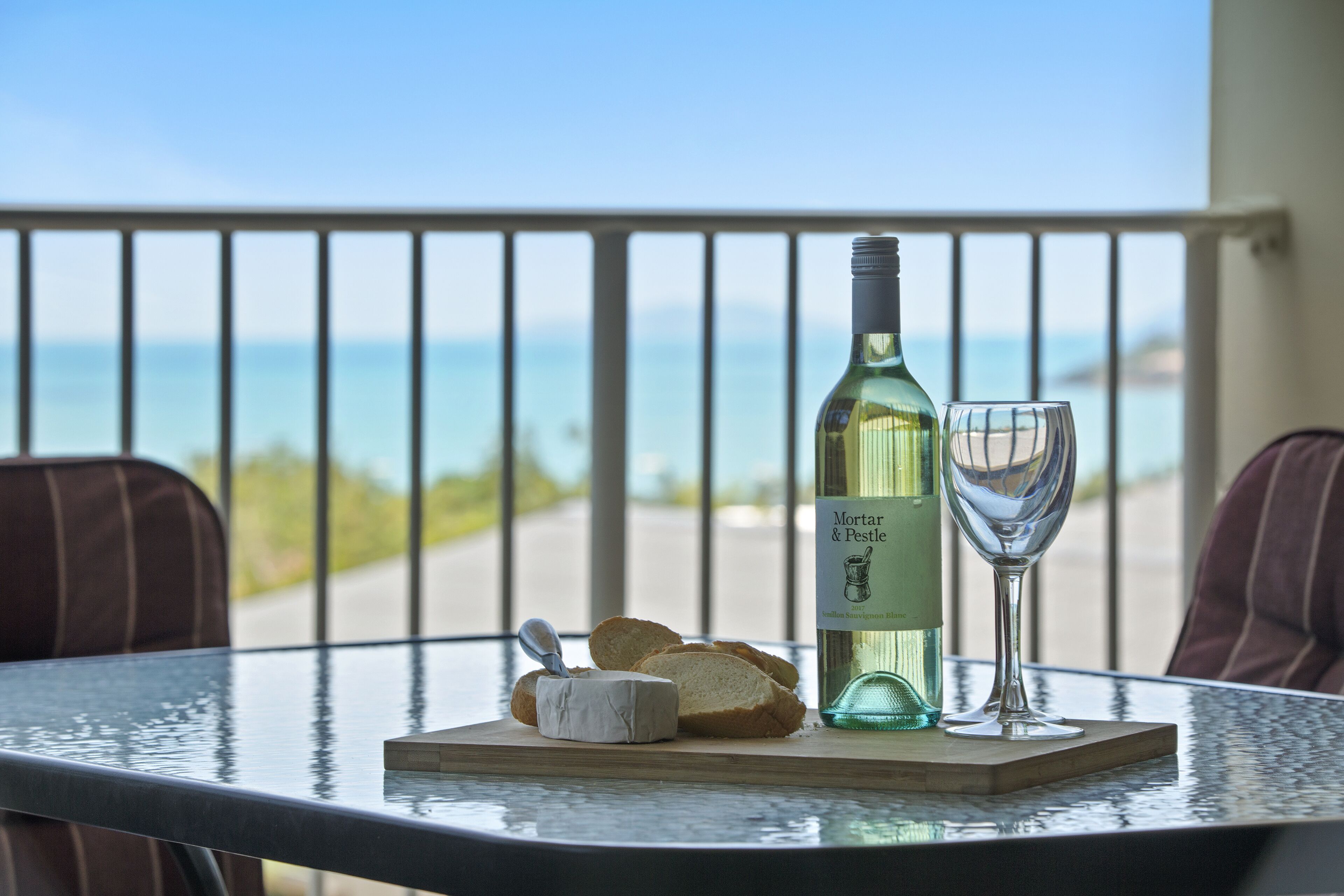 Et bord med en vinflaske, et glas og ost på en balkon med udsigt over havet.