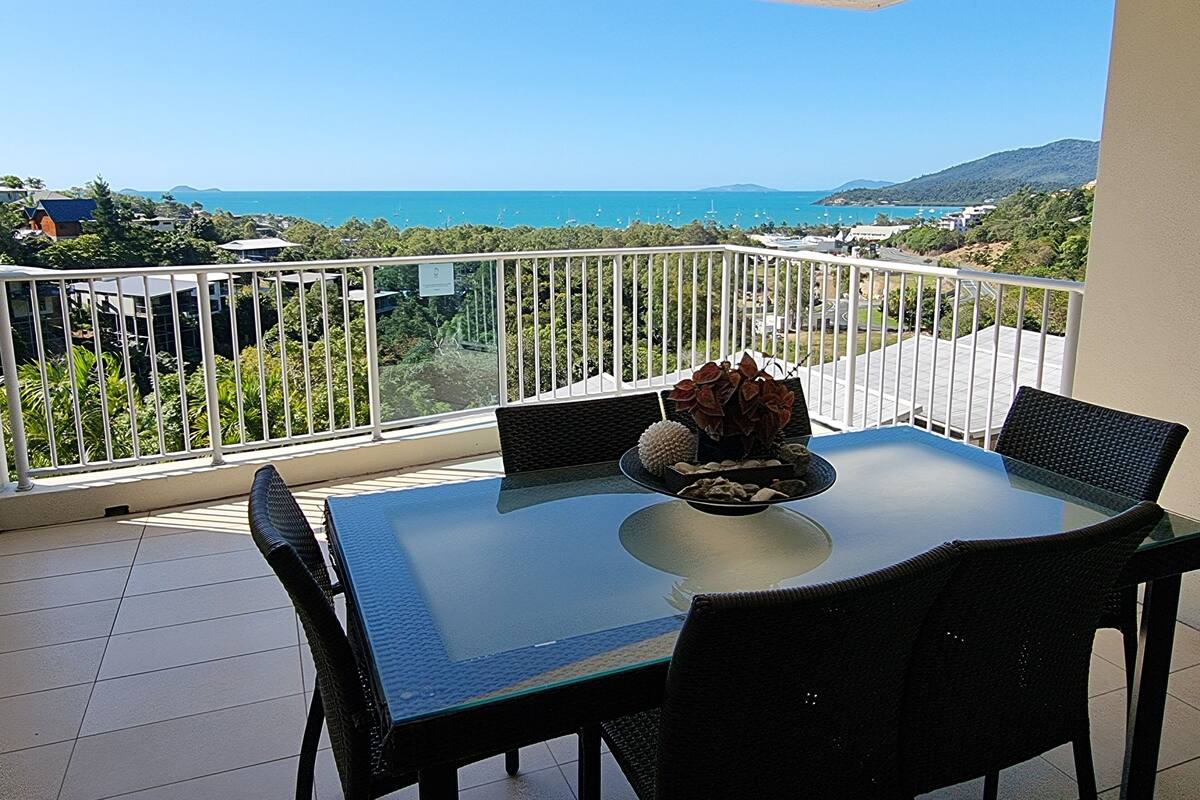Un balcon avec une table et des chaises en verre, une plante en pot et une vue sur la mer et la verdure.