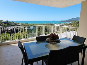 Een balkon met een glazen tafel en stoelen, een bloempot en uitzicht op zee en groen.