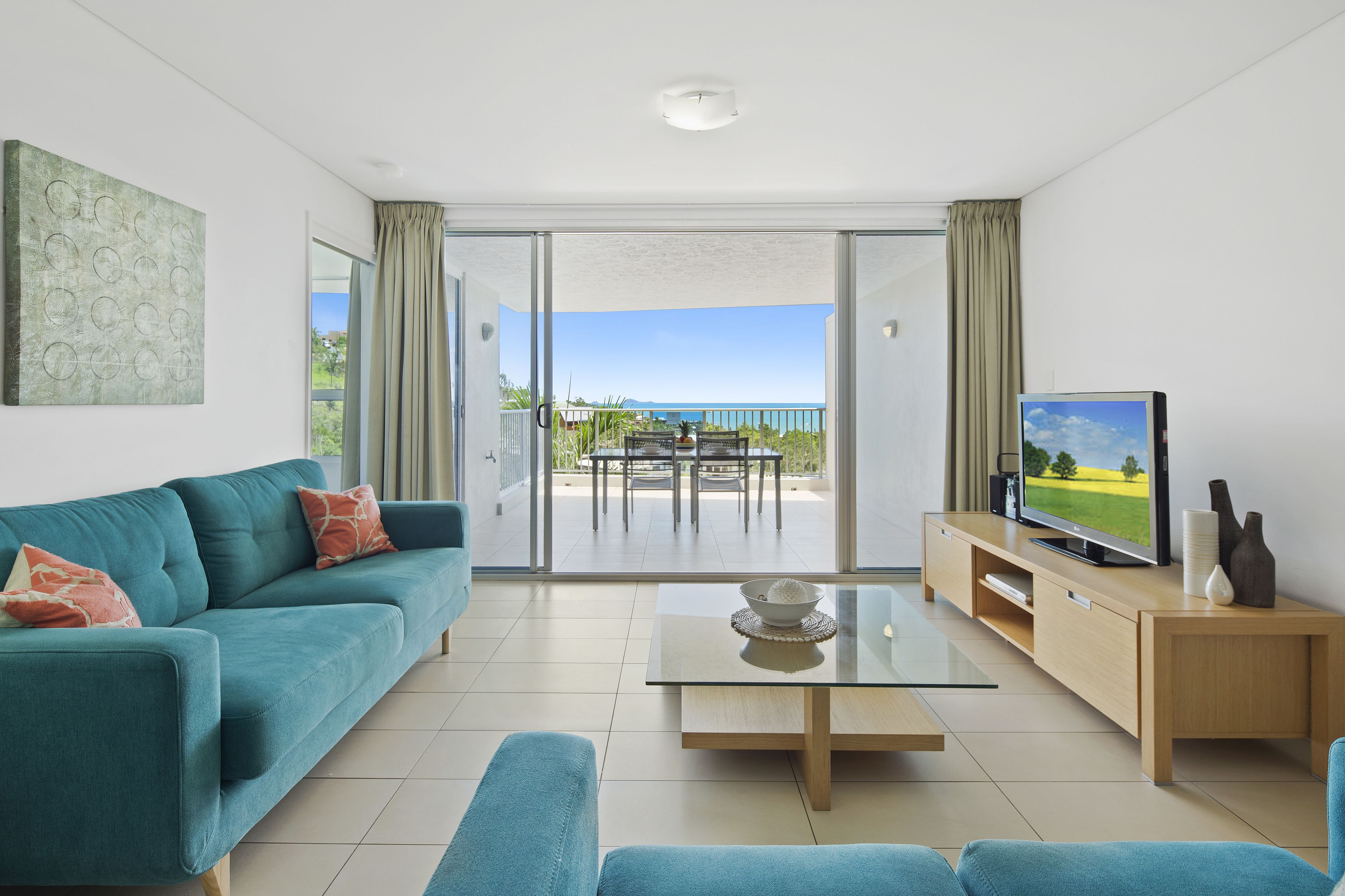 A modern living room with a sofa, a coffee table, a TV on a wooden stand, and a balcony with a view of the sea.