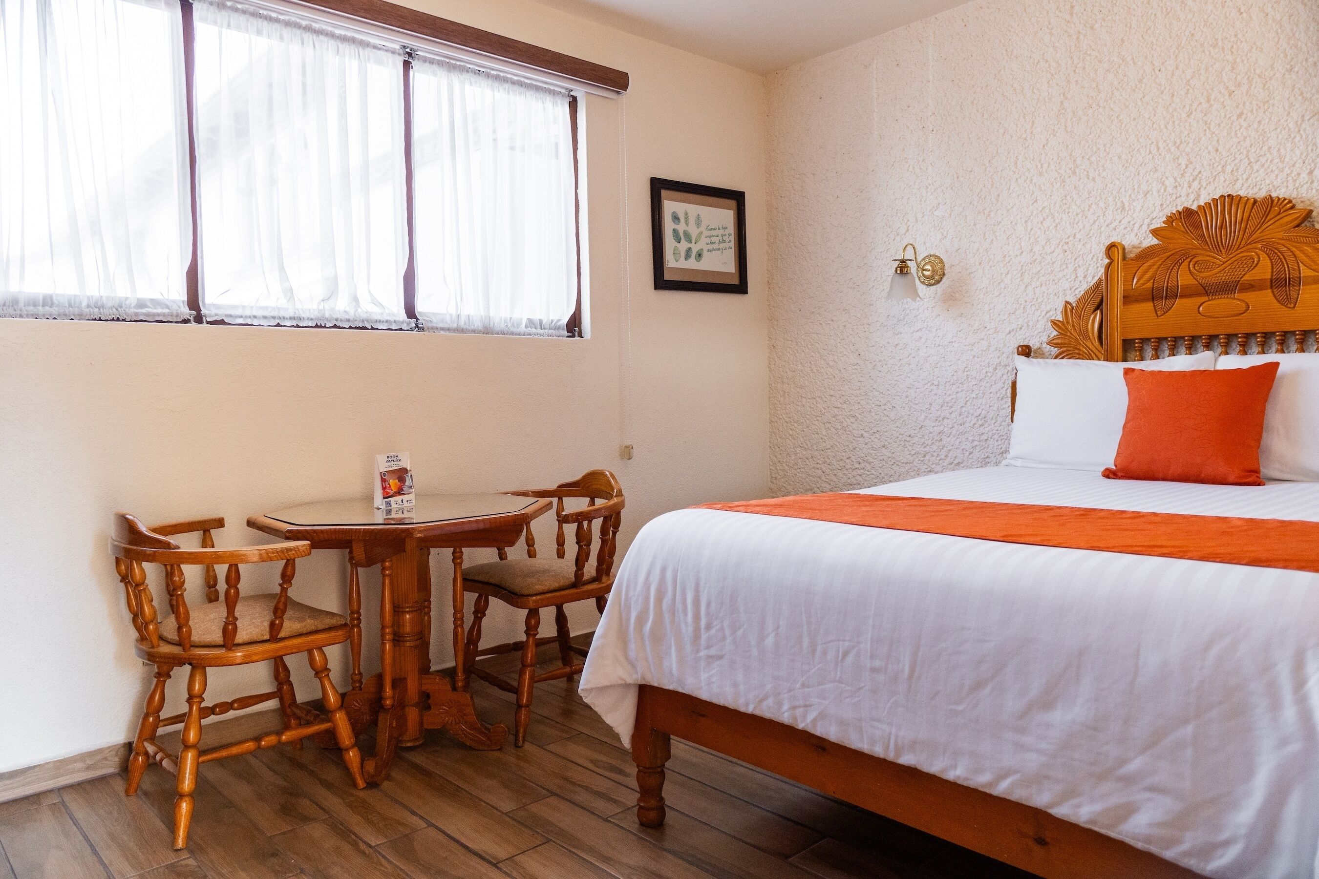 A bedroom with a wooden bed, white bedding, a small wooden table with chairs, and a window with sheer curtains.