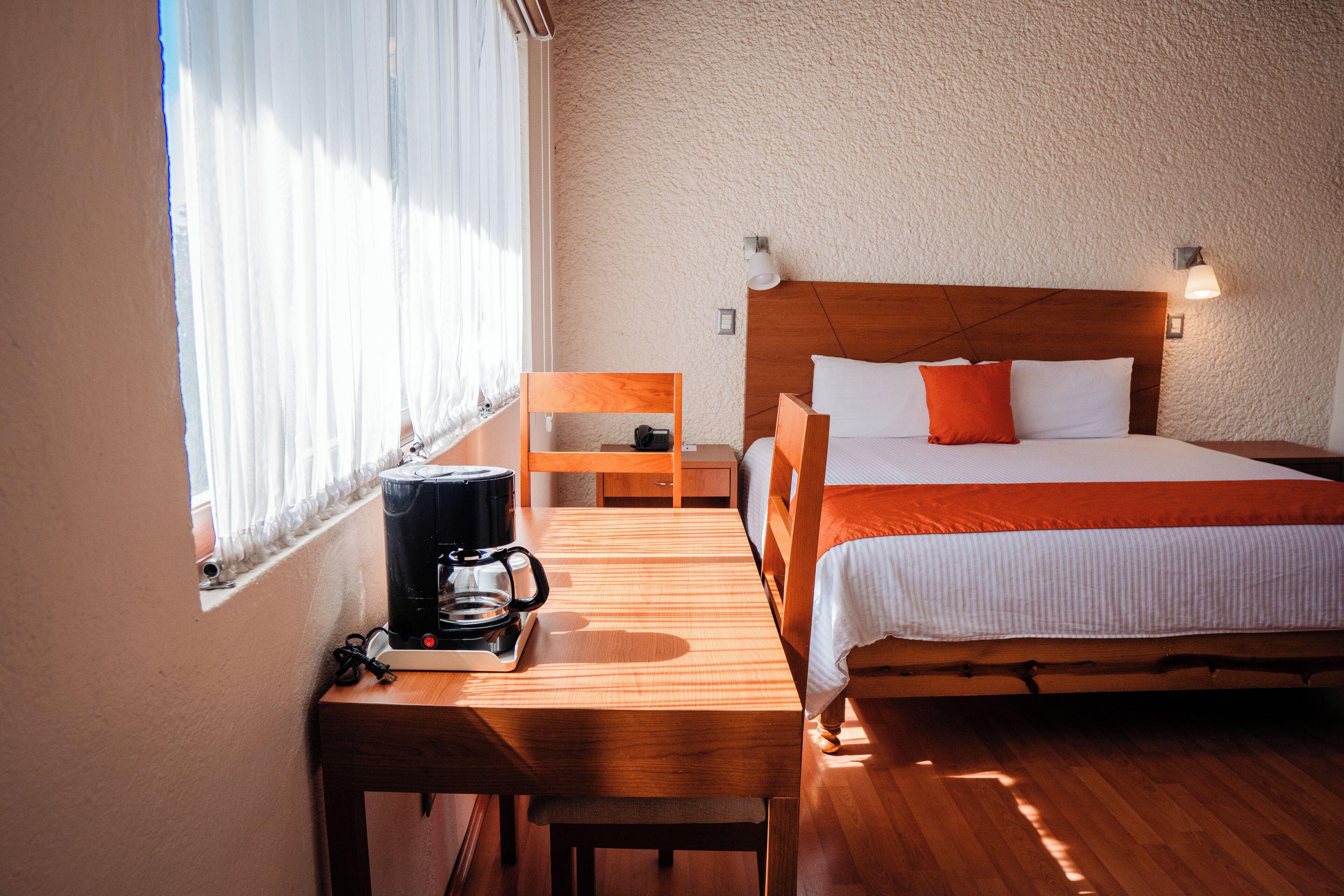 Habitación de hotel con cama de madera, un escritorio de madera con cafetera, una silla y una ventana con cortinas traslúcidas.