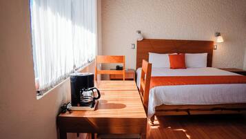 Habitación de hotel con cama de madera, un escritorio de madera con cafetera, una silla y una ventana con cortinas traslúcidas.