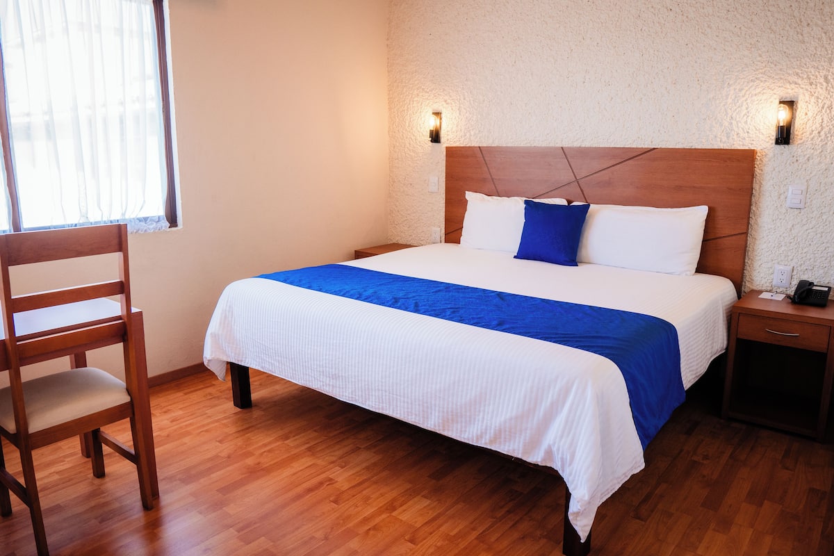 A neatly made bed with a wooden headboard, a bedside table with a phone, and a chair.