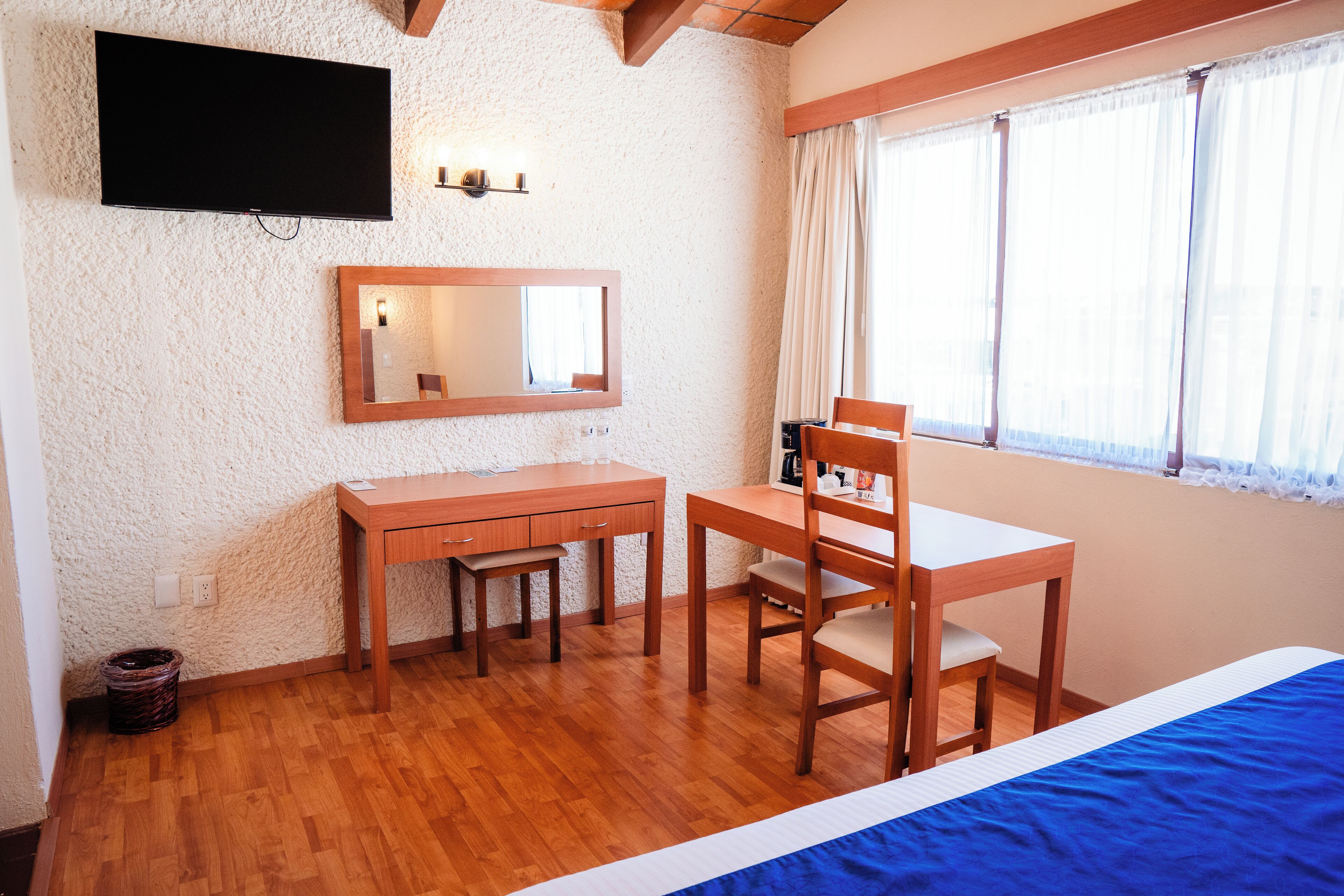 A room with a bed, a desk with a chair, a television mounted on the wall, and a window with curtains.