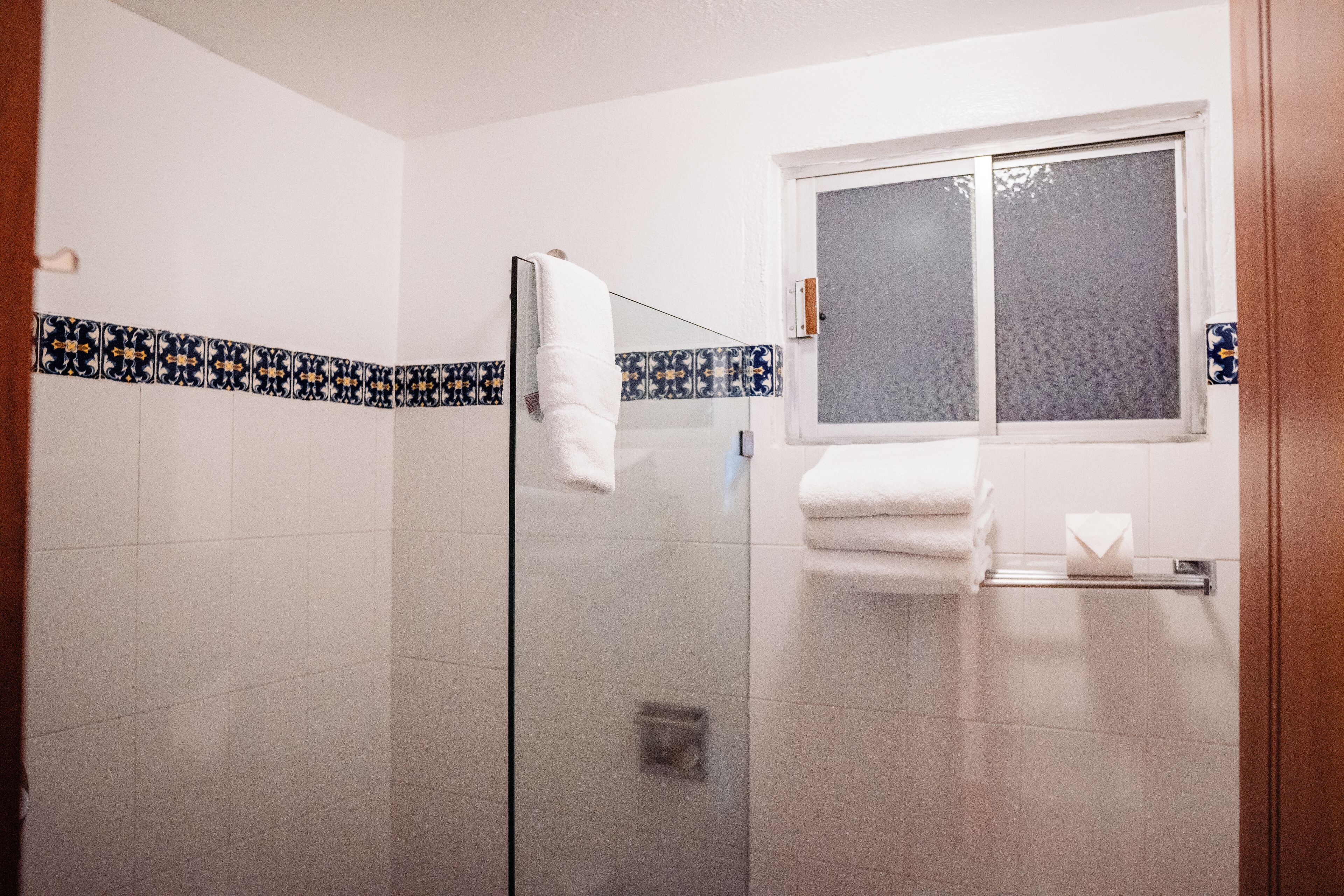 Un baño con cabina de ducha de vidrio, paredes cubiertas de azulejos blancos, una ventana y un perchero con toallas dobladas.
