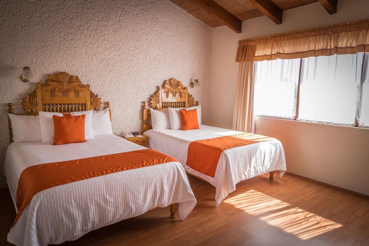 A room with two beds, wooden headboards, white bedding, orange accents, a wooden floor, a window with curtains, and a textured wall.
