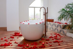 Una bañera grande llena de pétalos de rosa, suelo de madera y una planta en maceta.