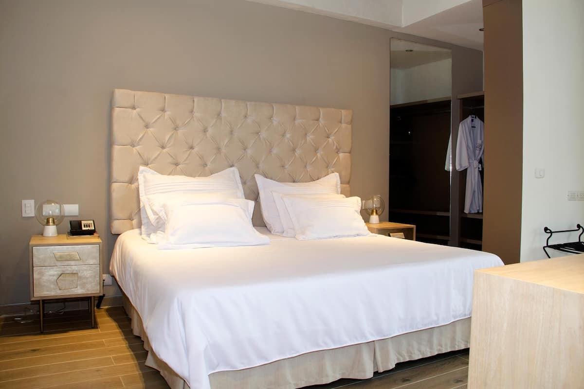 A bedroom with a tufted headboard, a bed with white linens, a bedside table with a lamp, and a wooden floor.
