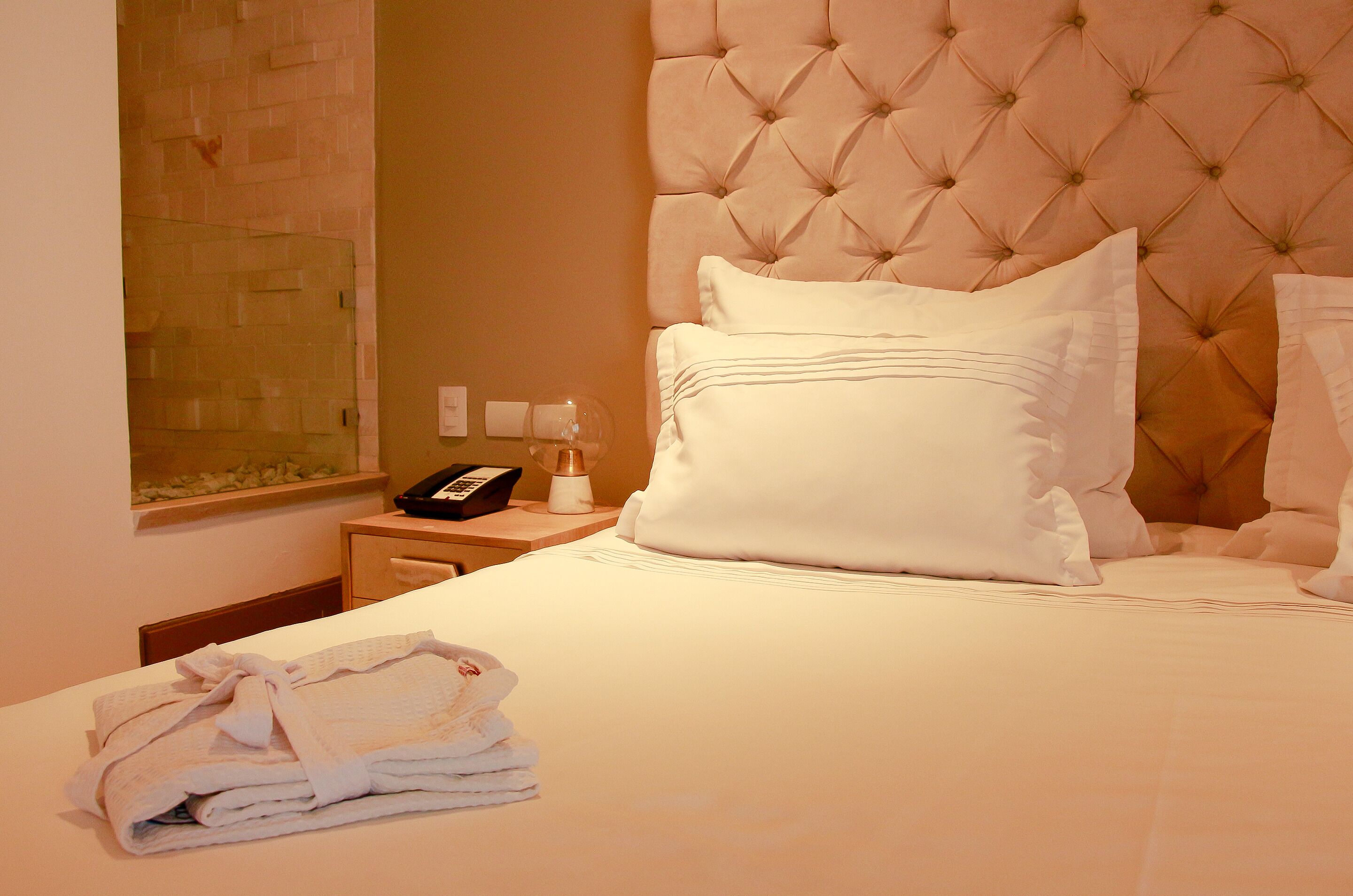 Una habitación de hotel con un cabecero acolchado, una cama bien hecha con ropa blanca, una mesita de noche con lámpara y una toalla doblada en el suelo.