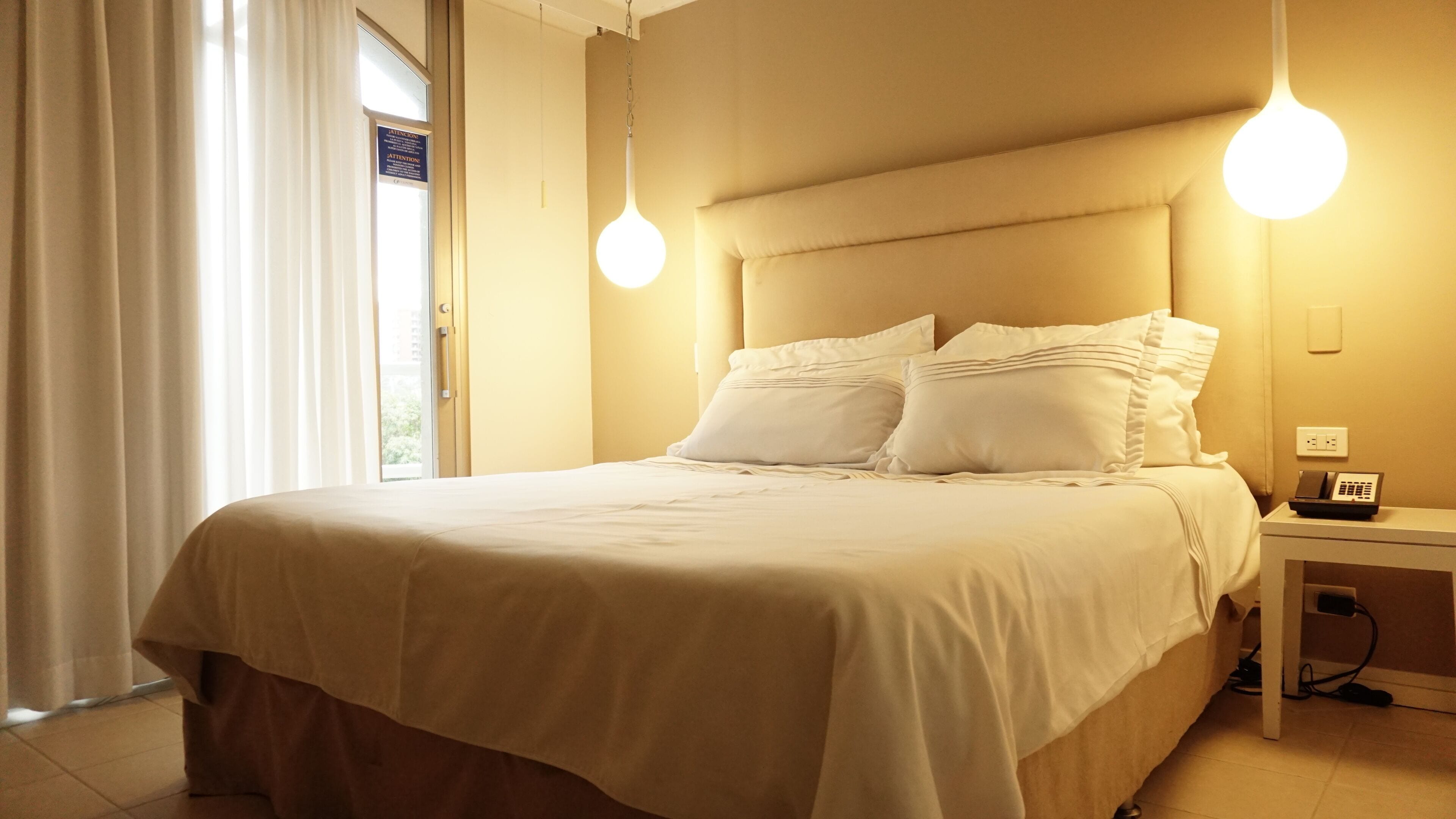 Une chambre d’hôtel avec un grand lit, deux lampes de chevet, un téléphone sur la table de chevet et une vue sur l’extérieur par une fenêtre munie de rideaux.