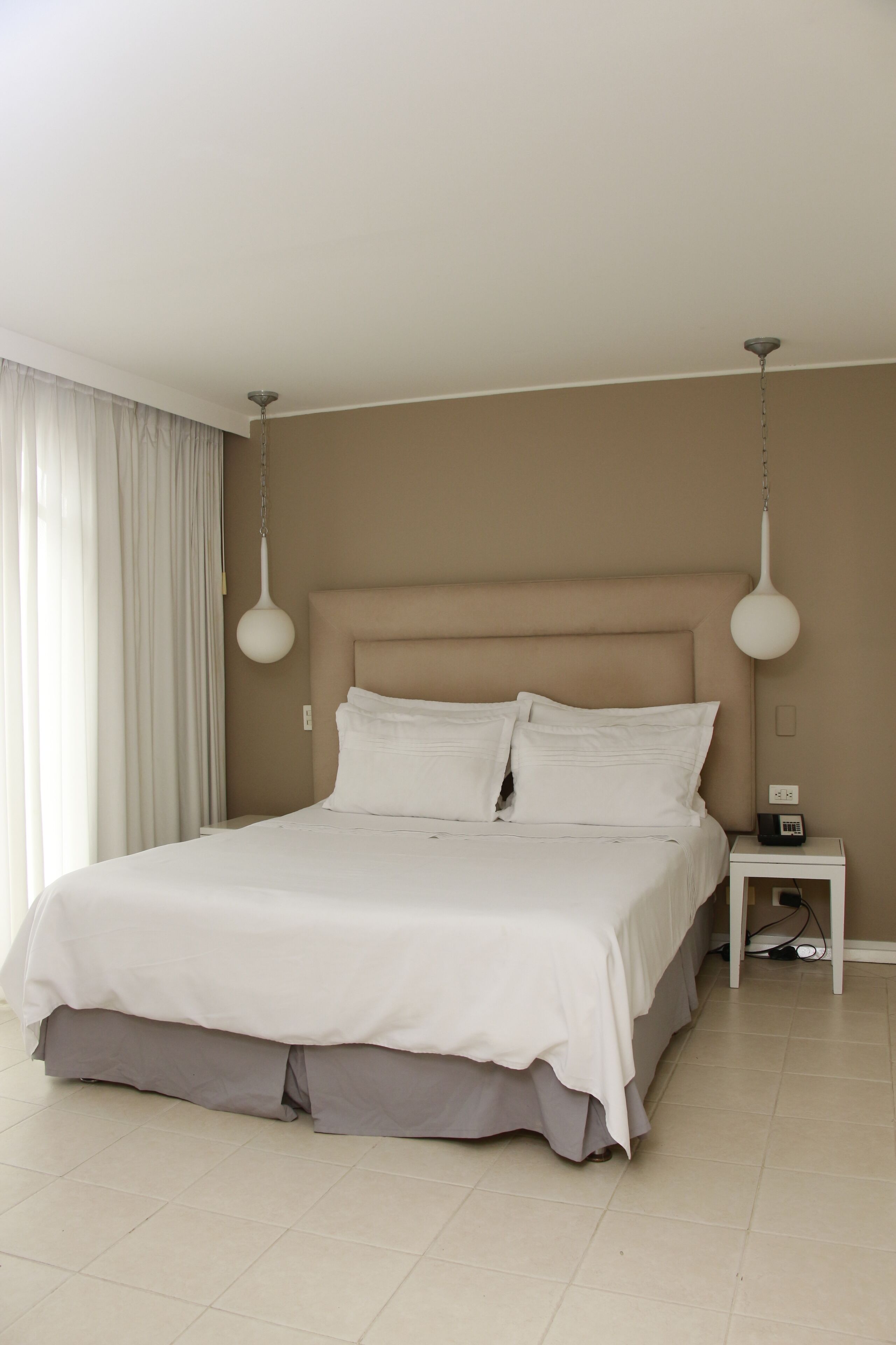 A neatly made bed with white linens, a bedside table with a lamp, and a view of a window with curtains.