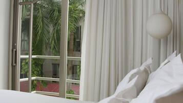 A hotel room with a large bed, a view through a sliding glass door to a balcony with greenery, and a hanging light fixture.