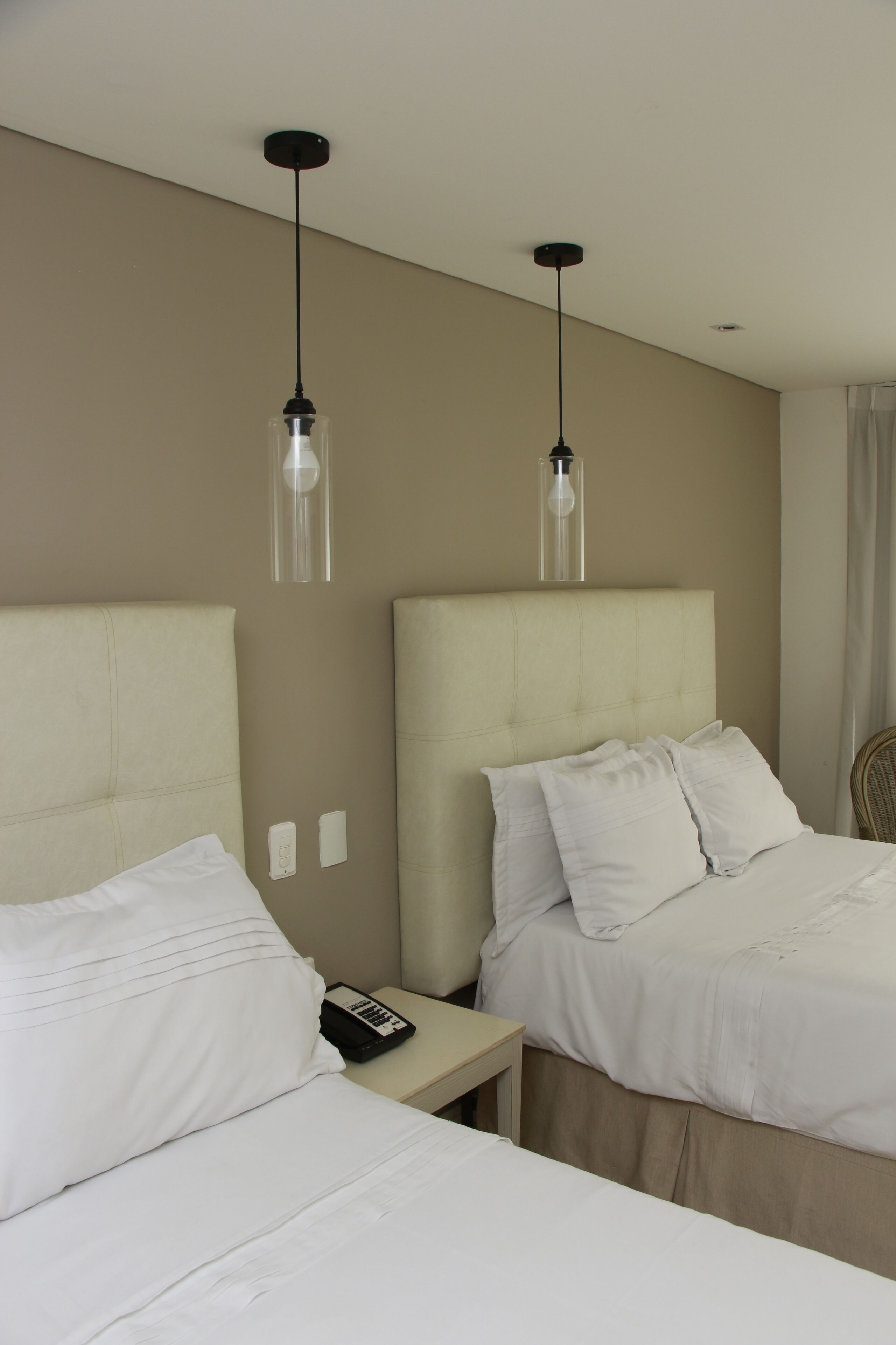 Een hotelkamer met twee bedden, elk met hoofdeinde en wit beddengoed. Twee hanglampen hangen aan het plafond, en een klein nachtkastje met een telefoon en een boek.