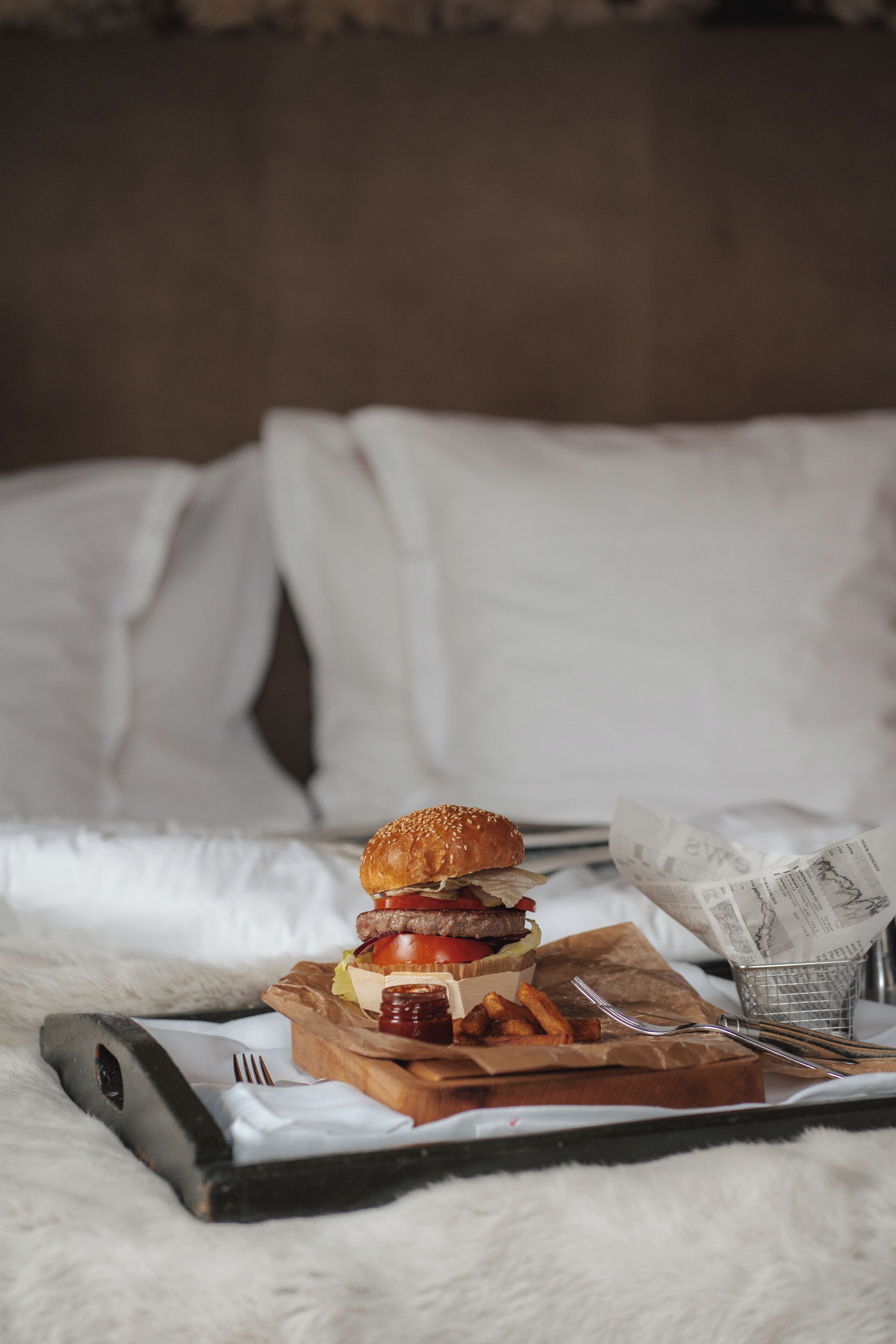 Un lit avec une tête de lit en bois, un oreiller blanc, une couverture et un repas composé d’un hamburger, de frites et d’une boisson, le tout présenté sur un plateau.