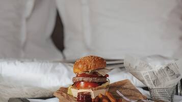 Un lit avec une tête de lit en bois, un oreiller blanc, une couverture et un repas composé d’un hamburger, de frites et d’une boisson, le tout présenté sur un plateau.