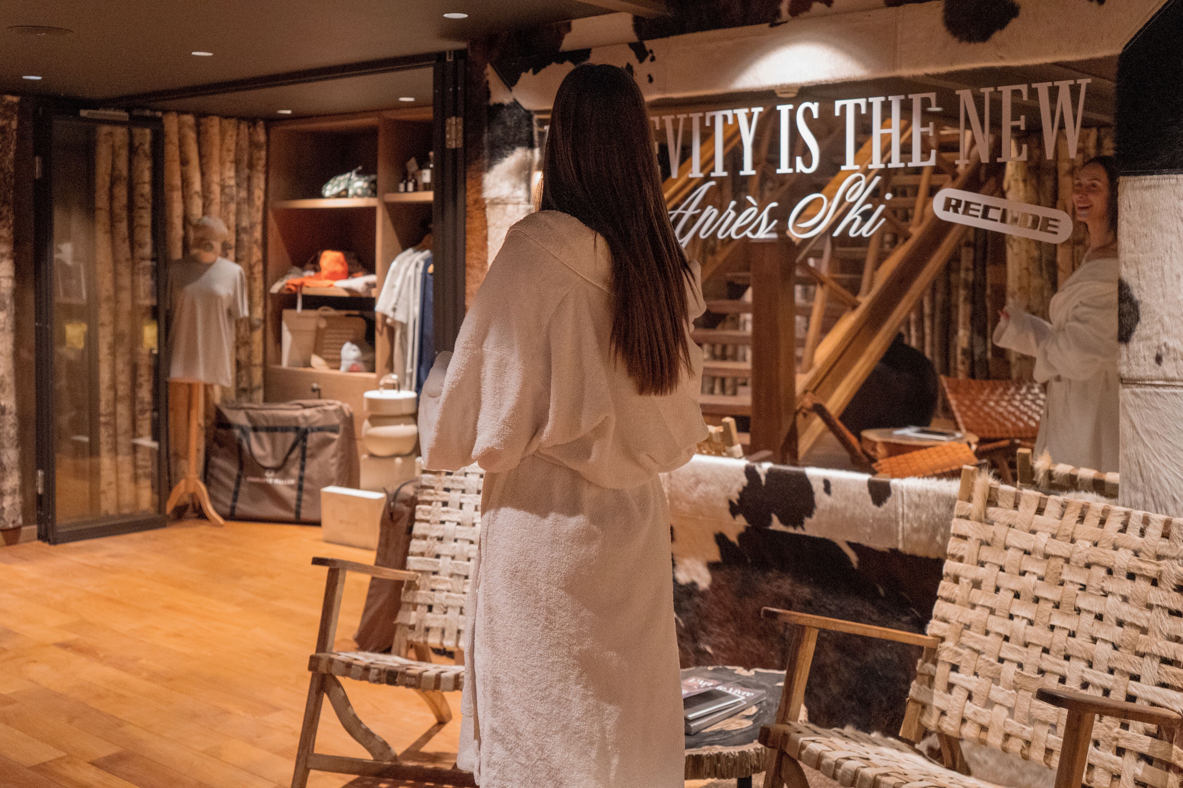 A person in a white robe standing in a room with wooden floors and a cowhide wall. There are wicker chairs and a wooden staircase.
