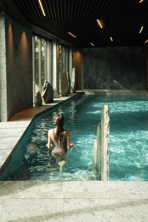 A pool area with a person sitting at the edge, large rocks, and a modern ceiling design. - Lodge Park (Megeve)