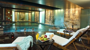 Un área de piscina interior moderna con sillones de descanso, una mesa con frutas y bebidas, y una pared de piedra.