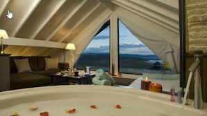 Habitación con una bañera grande llena de pétalos de rosa, vista a la playa y un rincón de descanso acogedor con una lámpara.