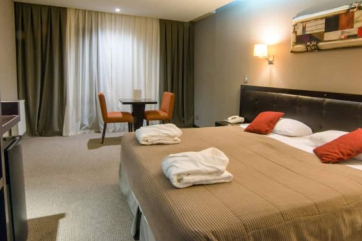 A hotel room with a large bed, two pillows, a bedside table, a lamp, a chair, and a TV.