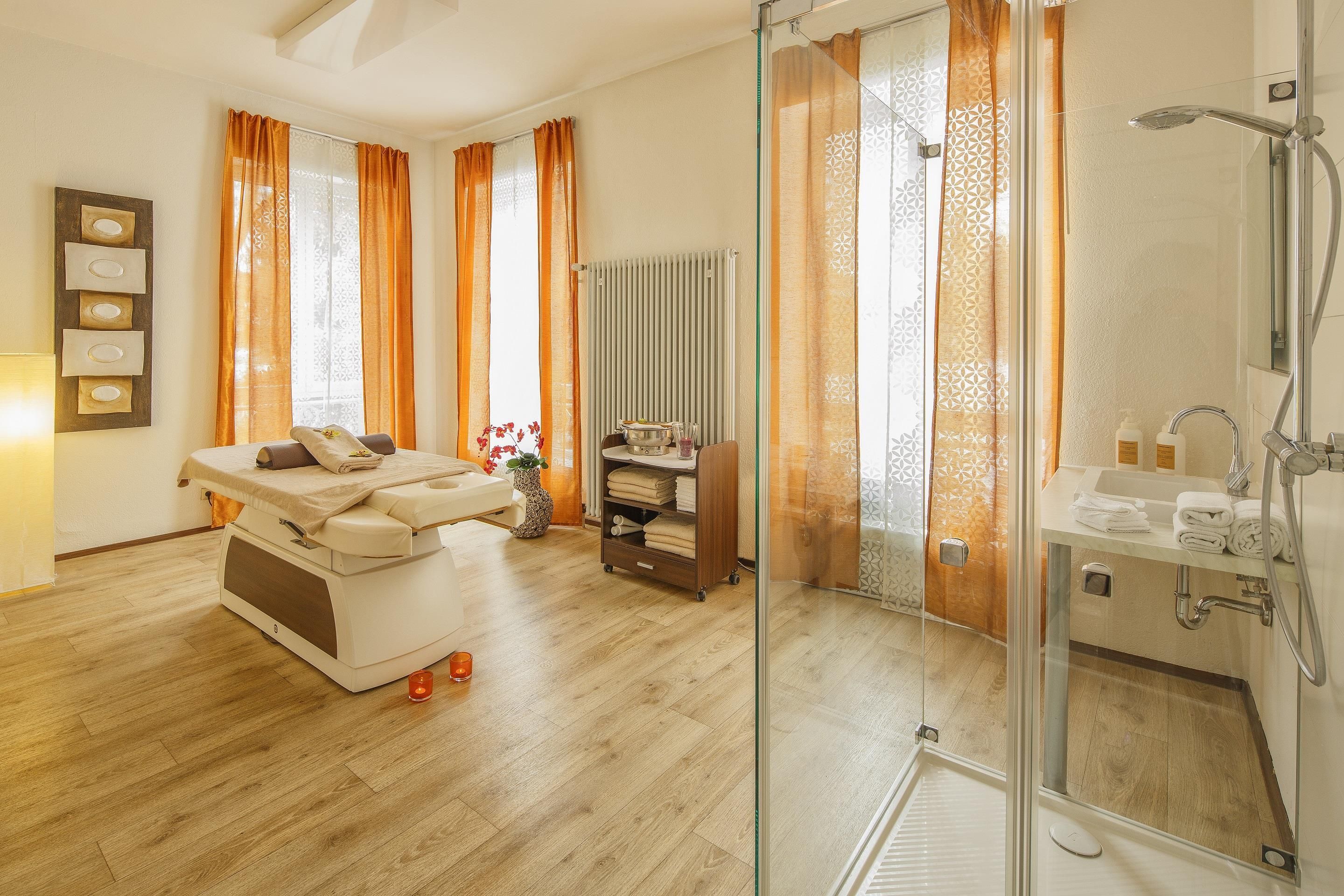 A spa room with a massage table, a small shelf with towels, and a shower area with a glass door.
