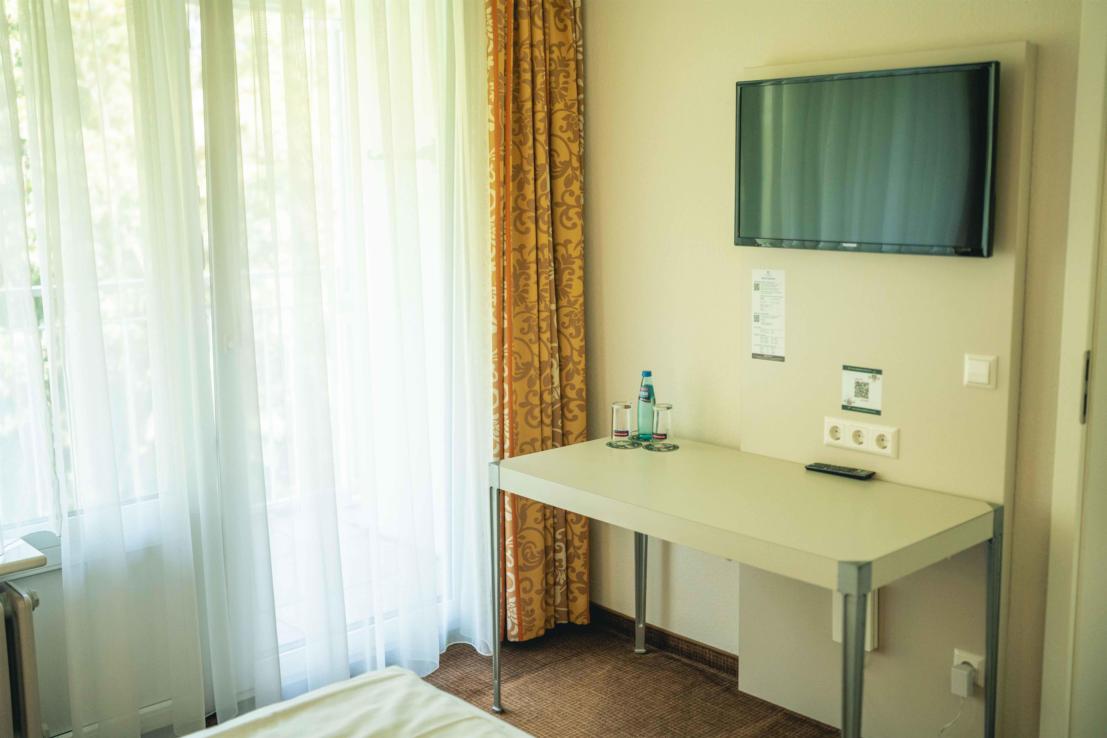 A hotel room with a bed, a desk, a television mounted on the wall, and a window with curtains.