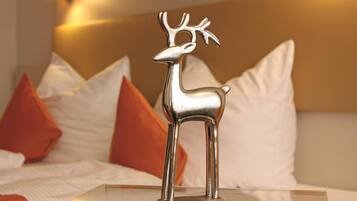 Eine silberne Hirschfigur steht auf einem klaren Glastisch in einem Hotelzimmer mit weißen und orangefarbenen Kissen.