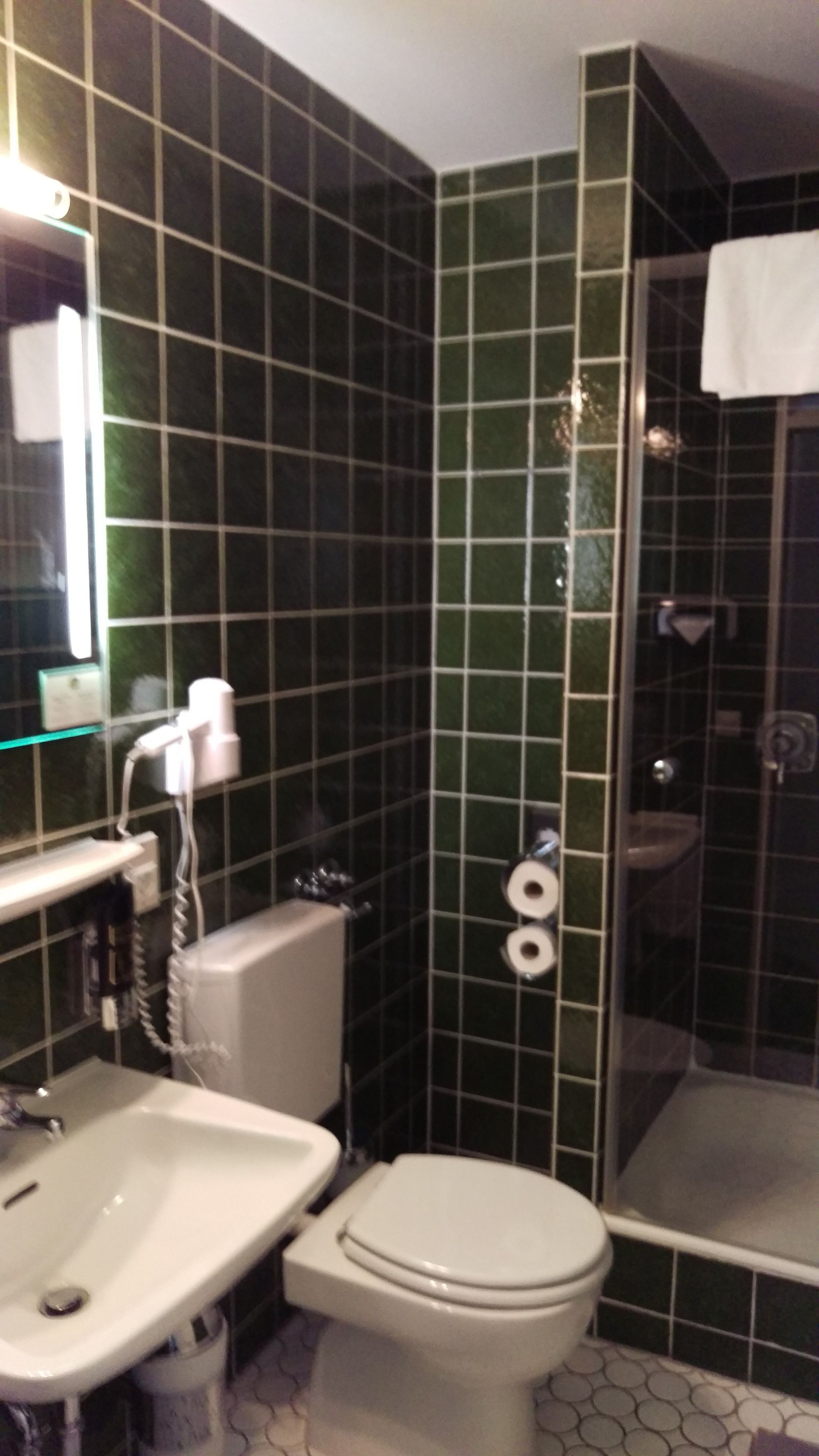 A bathroom with green tiled walls, a white sink, a toilet, and a glass shower enclosure.