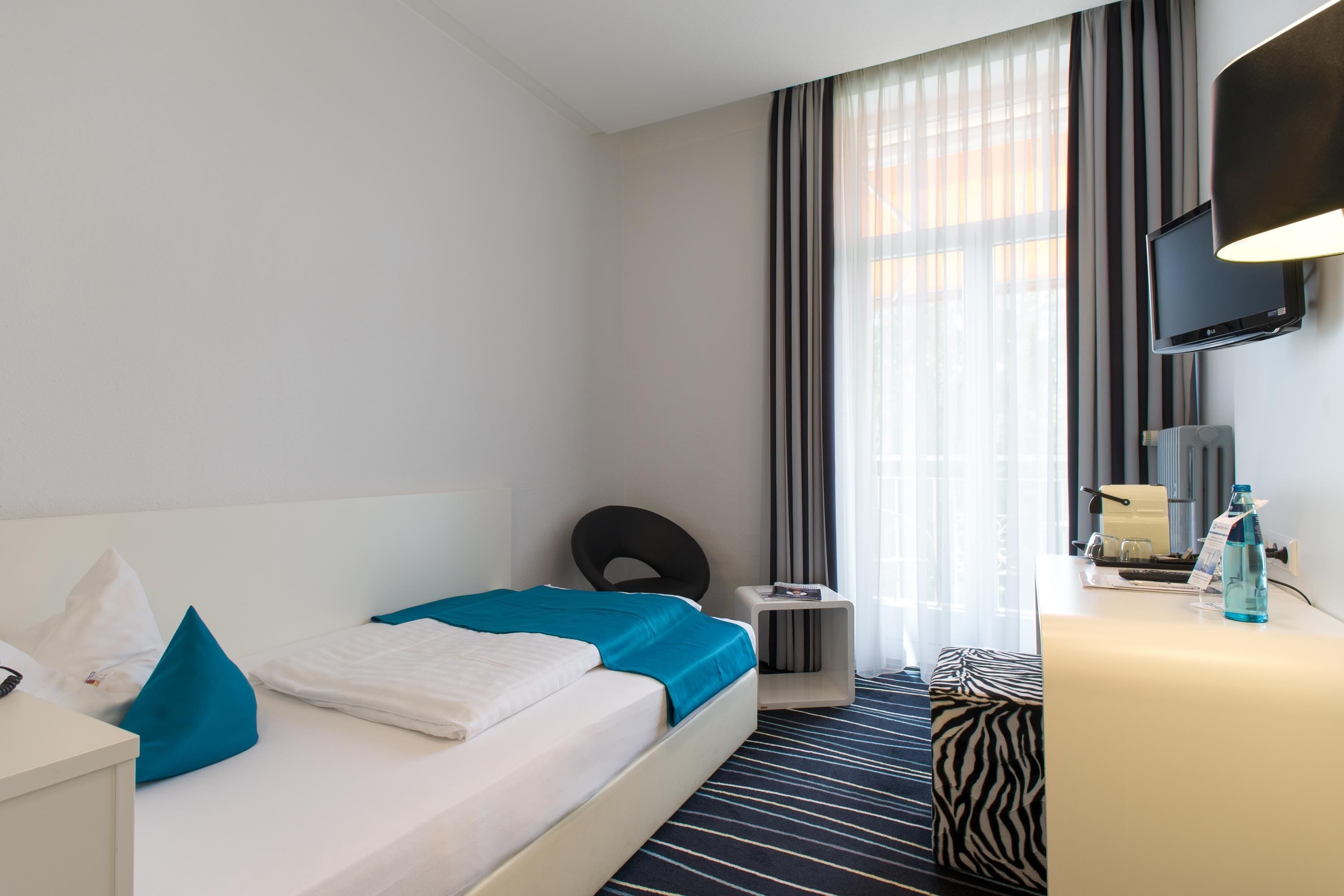 Habitación de hotel con cama, escritorio, silla, televisor y ventana con cortinas.