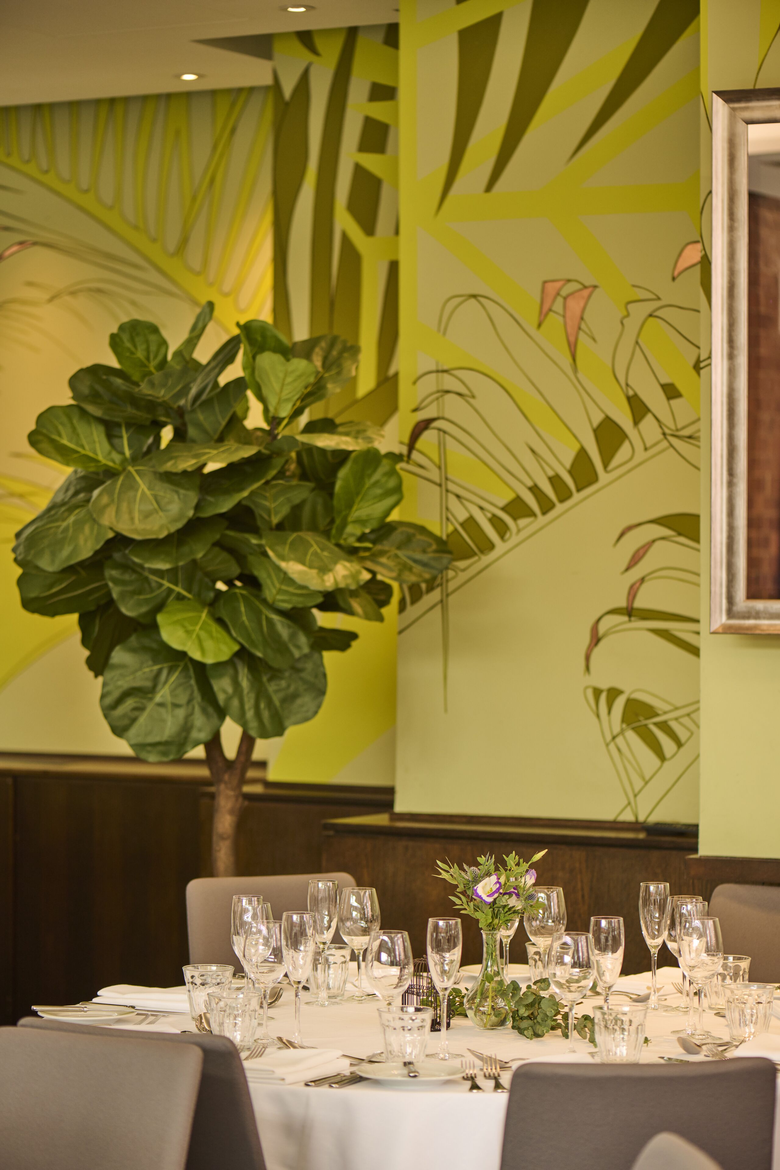 A dining table set for a meal with a large potted plant and a wall mural.