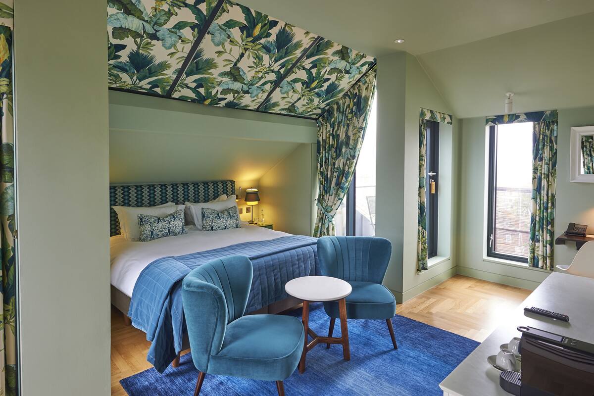 A bedroom with a large bed, two blue armchairs, a small round table, and a window with curtains.