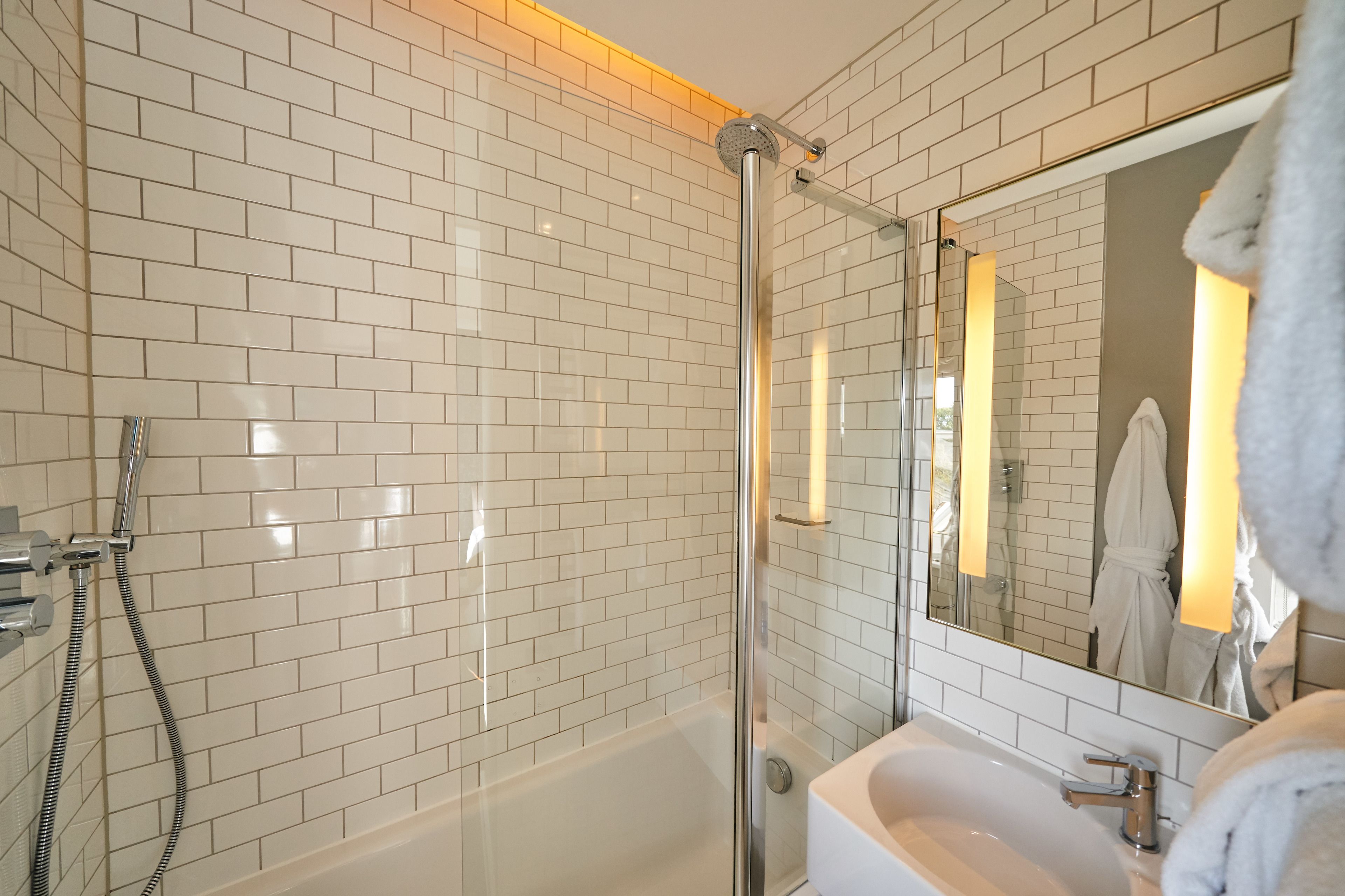 Une salle de bain moderne avec des carreaux de céramique blancs rectangulaires, une douche à parois de verre, un lavabo blanc et un miroir qui reflète une robe.