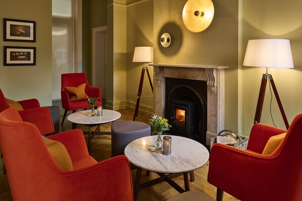 A cozy lounge area with red armchairs, a marble table, a fireplace, and two floor lamps.