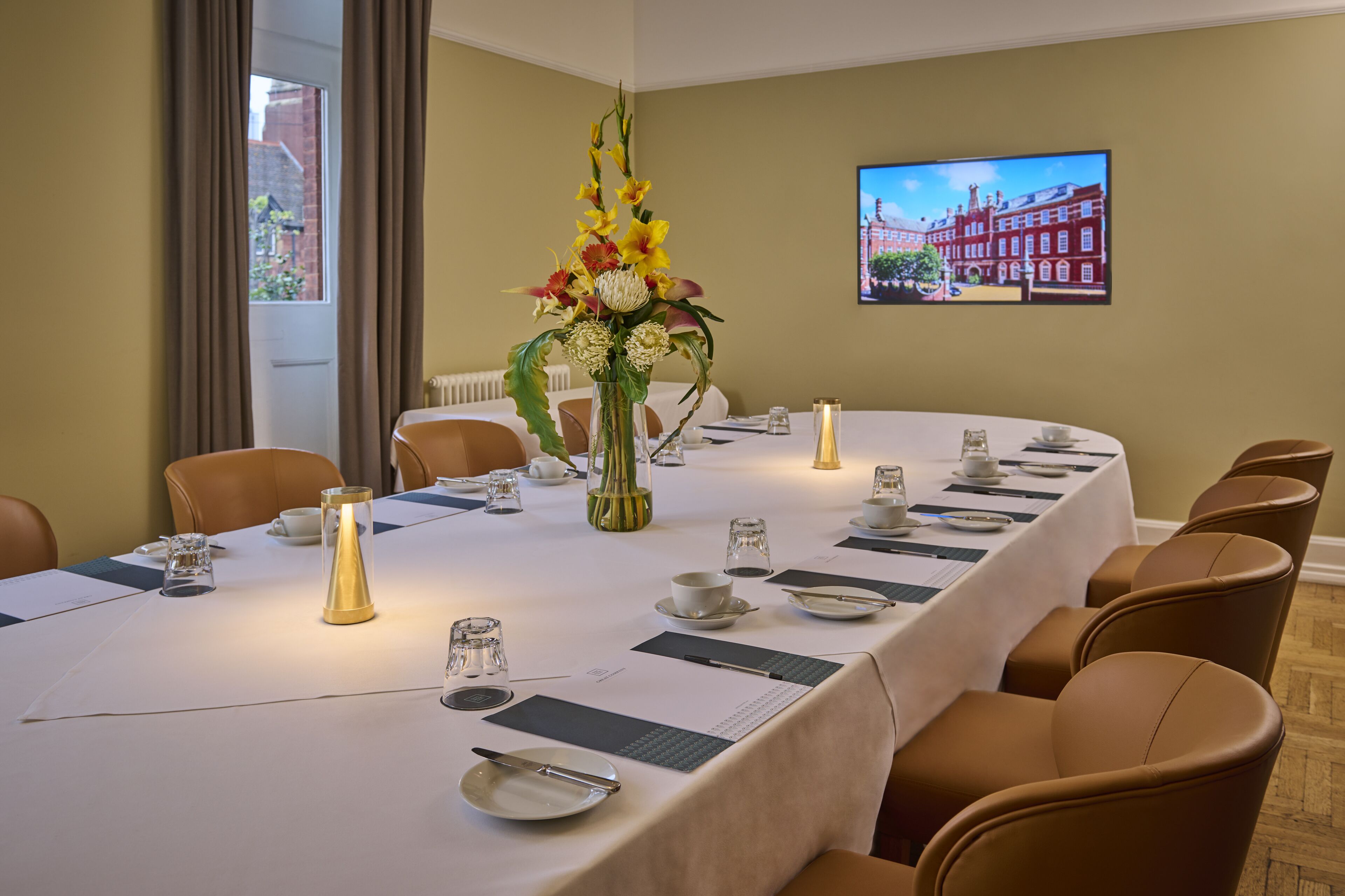 A conference room with a long table set for a meeting, equipped with chairs, a vase of flowers, and a television displaying a cityscape.