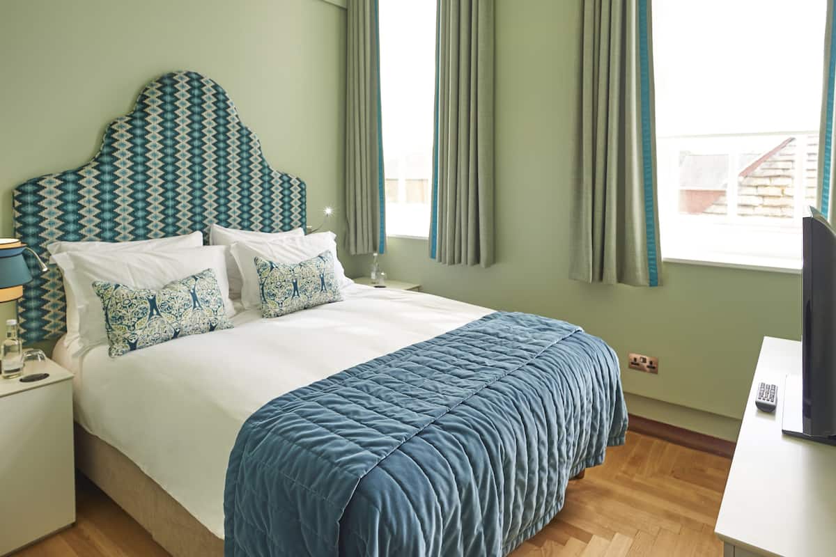 A neatly made bed with a blue quilt, a patterned headboard, and a bedside table with a lamp and a bottle.