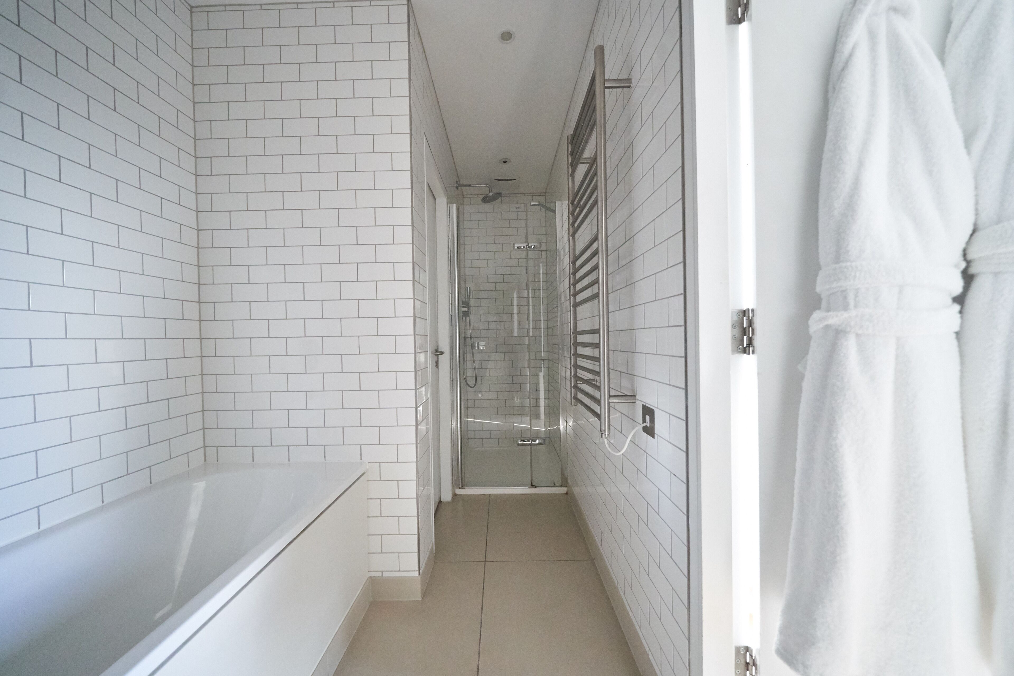 A narrow bathroom with white tiled walls, a bathtub, a glass-enclosed shower, and a towel rack.