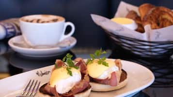 A breakfast plate with eggs benedict, a cup of coffee, and a basket of croissants.