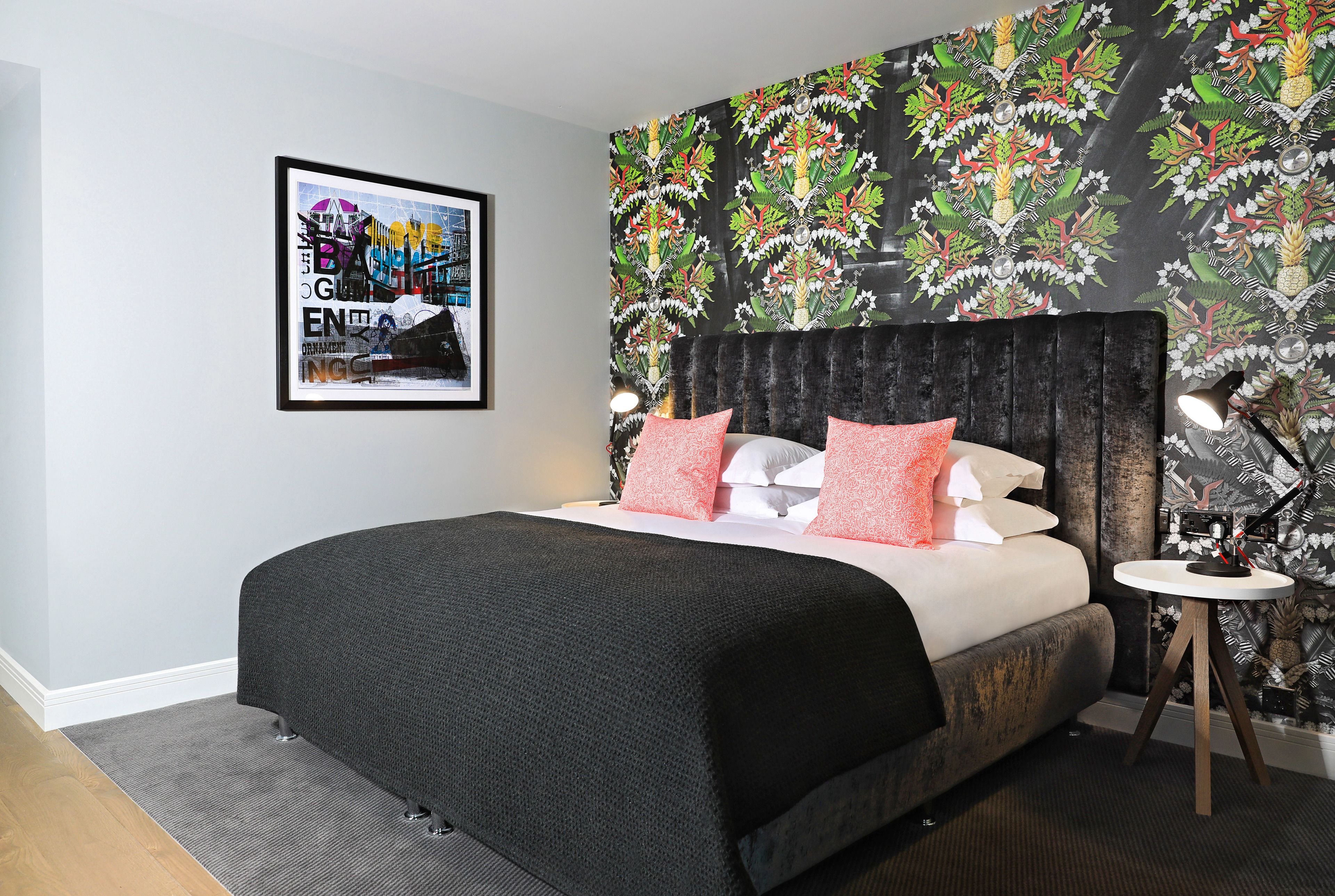 A modern bedroom with a large bed, a headboard, a side table, and a wall with a tropical wallpaper.