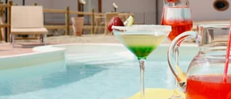 Zona de piscina con una silla de descanso, una mesa con bebidas y un edificio con ventanas arqueadas al fondo.