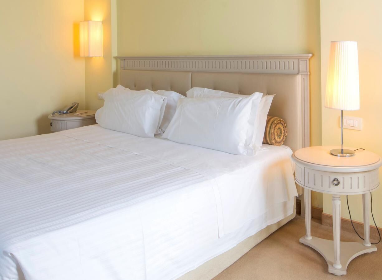 A neatly made bed with white linens, a round bedside table with a lamp, and a nightstand with a phone and a decorative item.