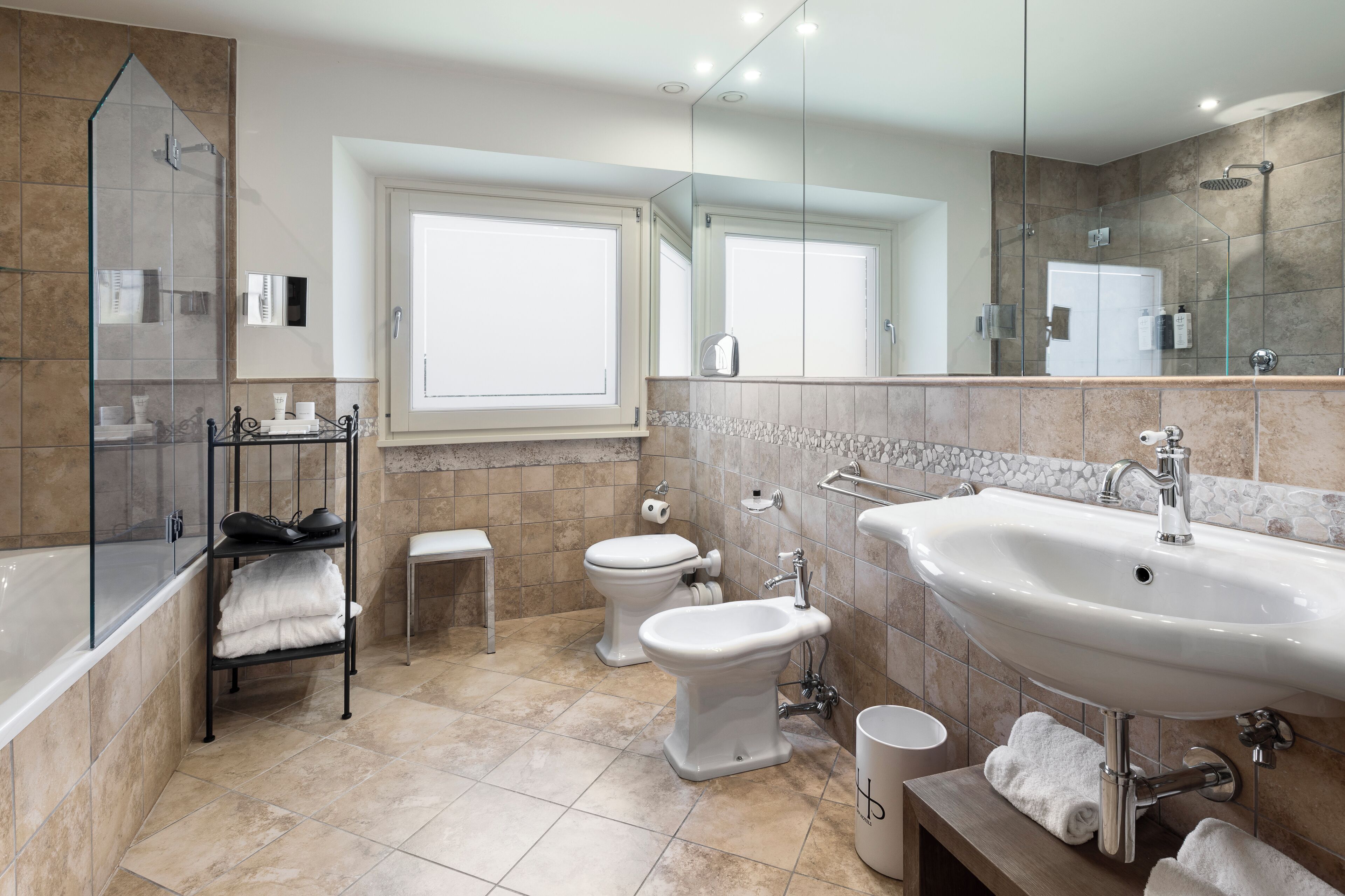 A modern bathroom with a bathtub, a large sink, a bidet, and a toilet. There is a window providing natural light and a mirror above the sink.