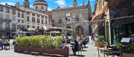 Un animado espacio al aire libre con un café, edificios de piedra, mesas exteriores y una jardinera grande con follaje.