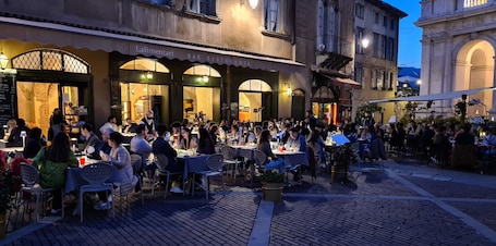 A bustling outdoor dining area with multiple tables and chairs, surrounded by buildings with arched windows and a stone facade.